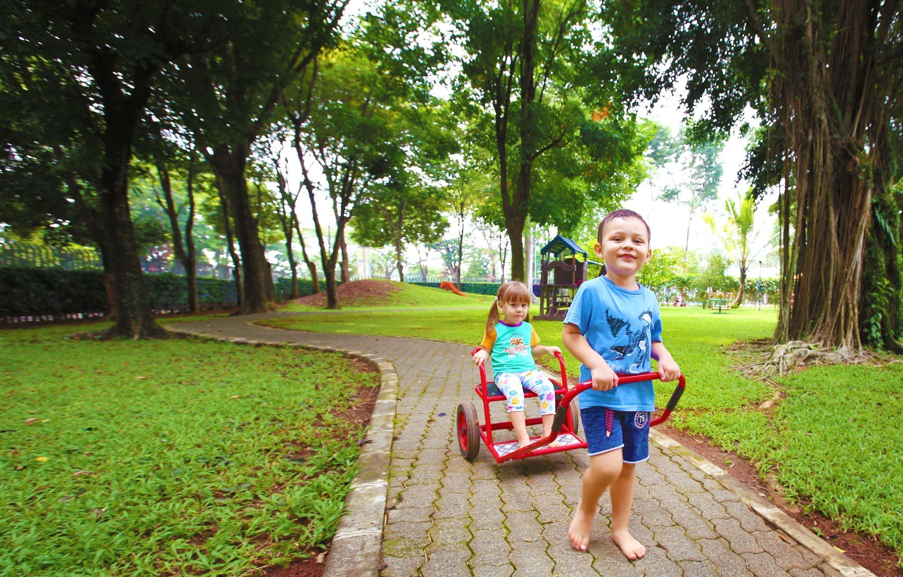 A boy is pulling a cart with two children in it in a park.