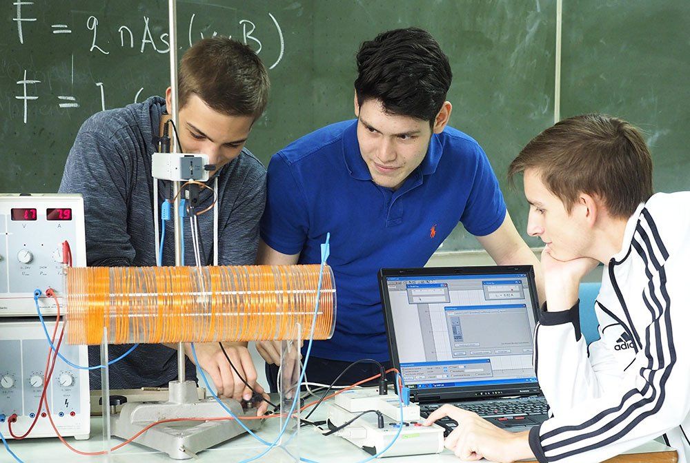 Three young men are working on a physics experiment in front of a blackboard.