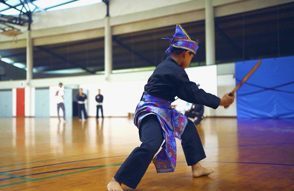 Child dressed in traditional martial arts attire practicing Silat in an indoor sports hall.