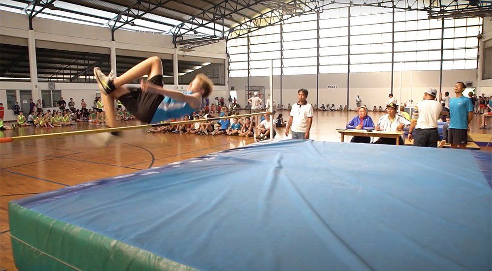 A person is jumping over a blue mat in a gym.