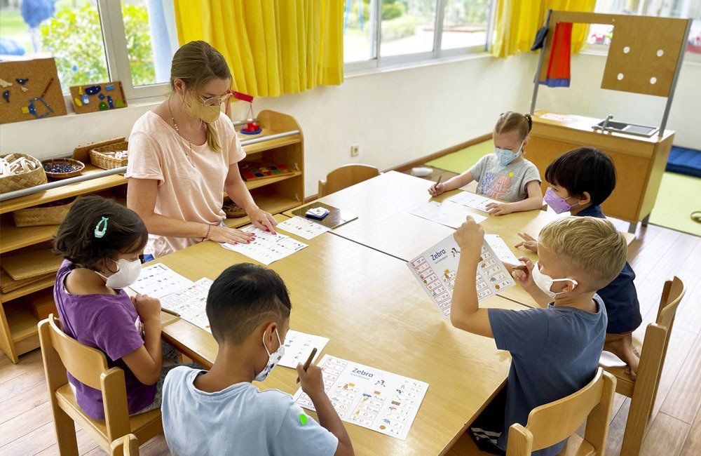 A group of children wearing face masks are sitting at a table with a teacher.