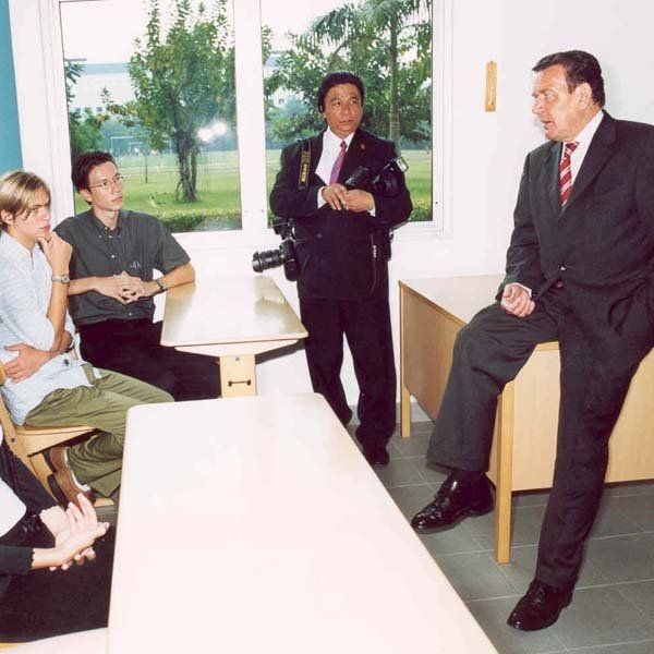 A man in a suit sits on a desk in front of a group of people