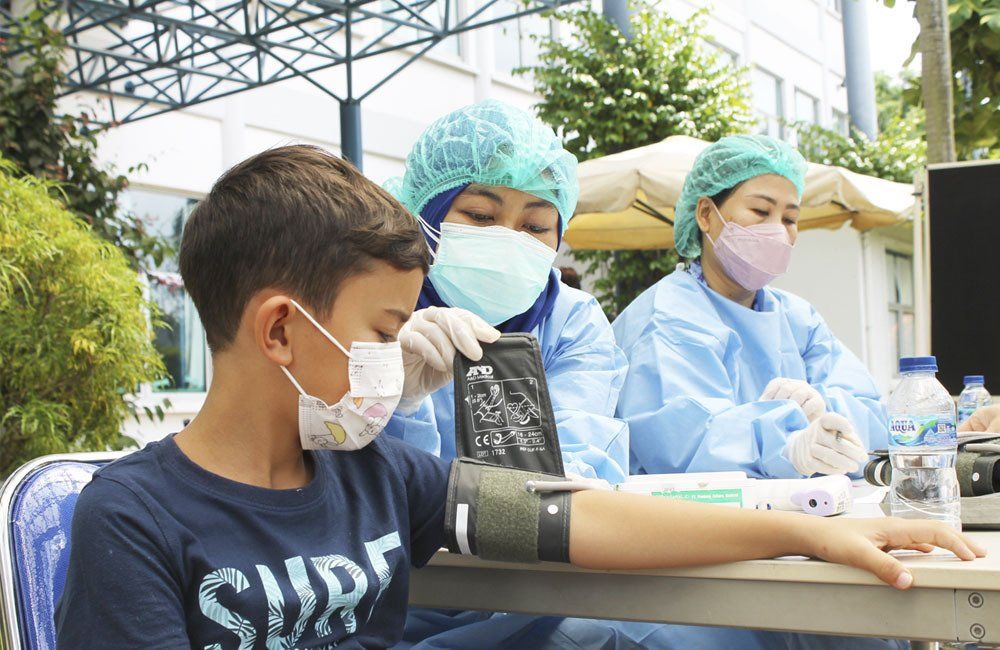 A young boy wearing a face mask while a healthcare worker in protective gear checks his blood pressure. 