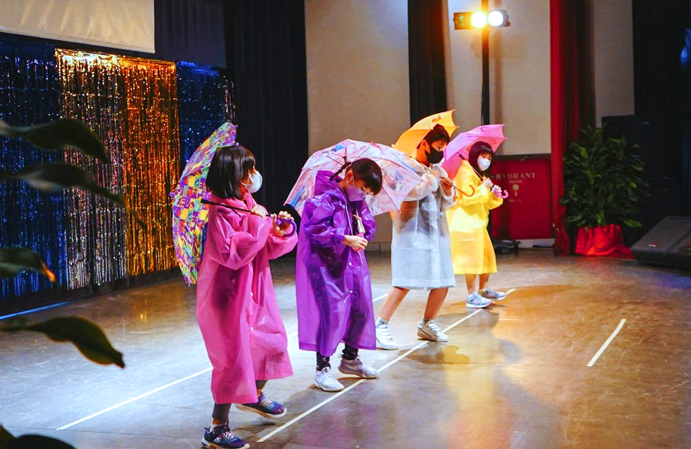 A group of children in raincoats and umbrellas are dancing on a stage.