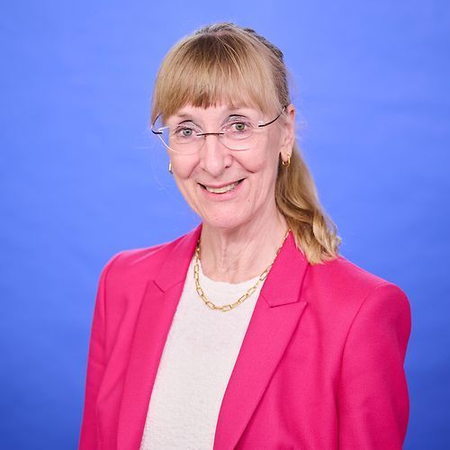 A woman wearing glasses and a pink jacket is smiling for the camera.