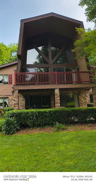 Two-story stone building with a large wooden balcony and covered roof, surrounded by trees and lawn