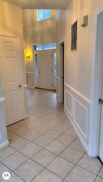 Bright hallway with white walls, tiled floor, front door, and wall sconces leading to a foyer.