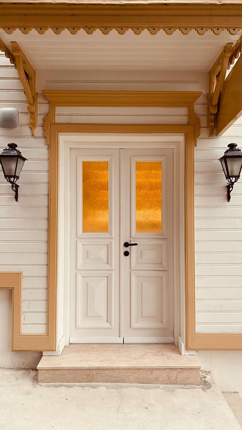 A person wearing a hat and vest uses a long-handled roller to paint a wall bright orange in a room with a wooden ladder.