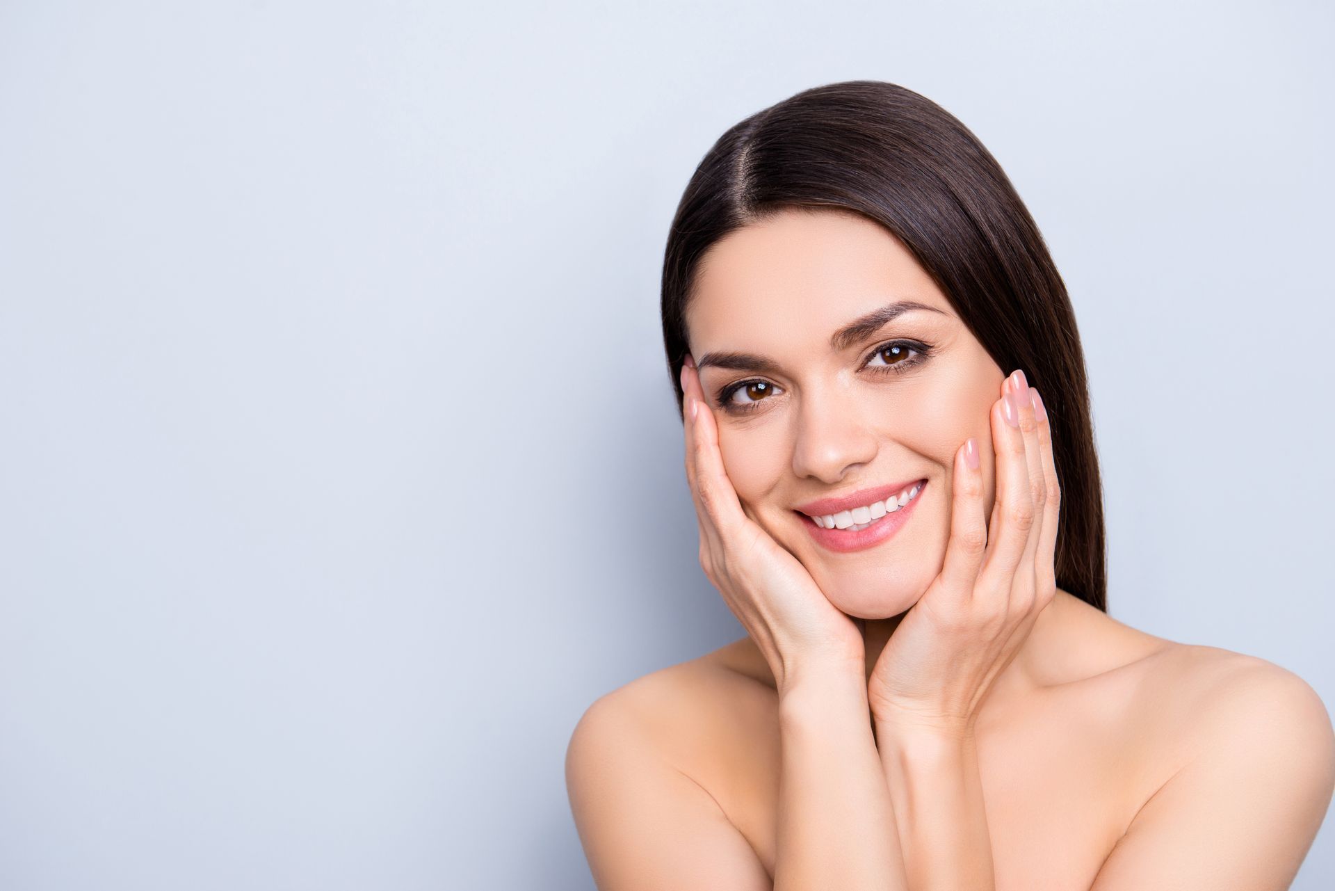 Woman with smooth skin, smiling, hands on cheeks. Light blue background.