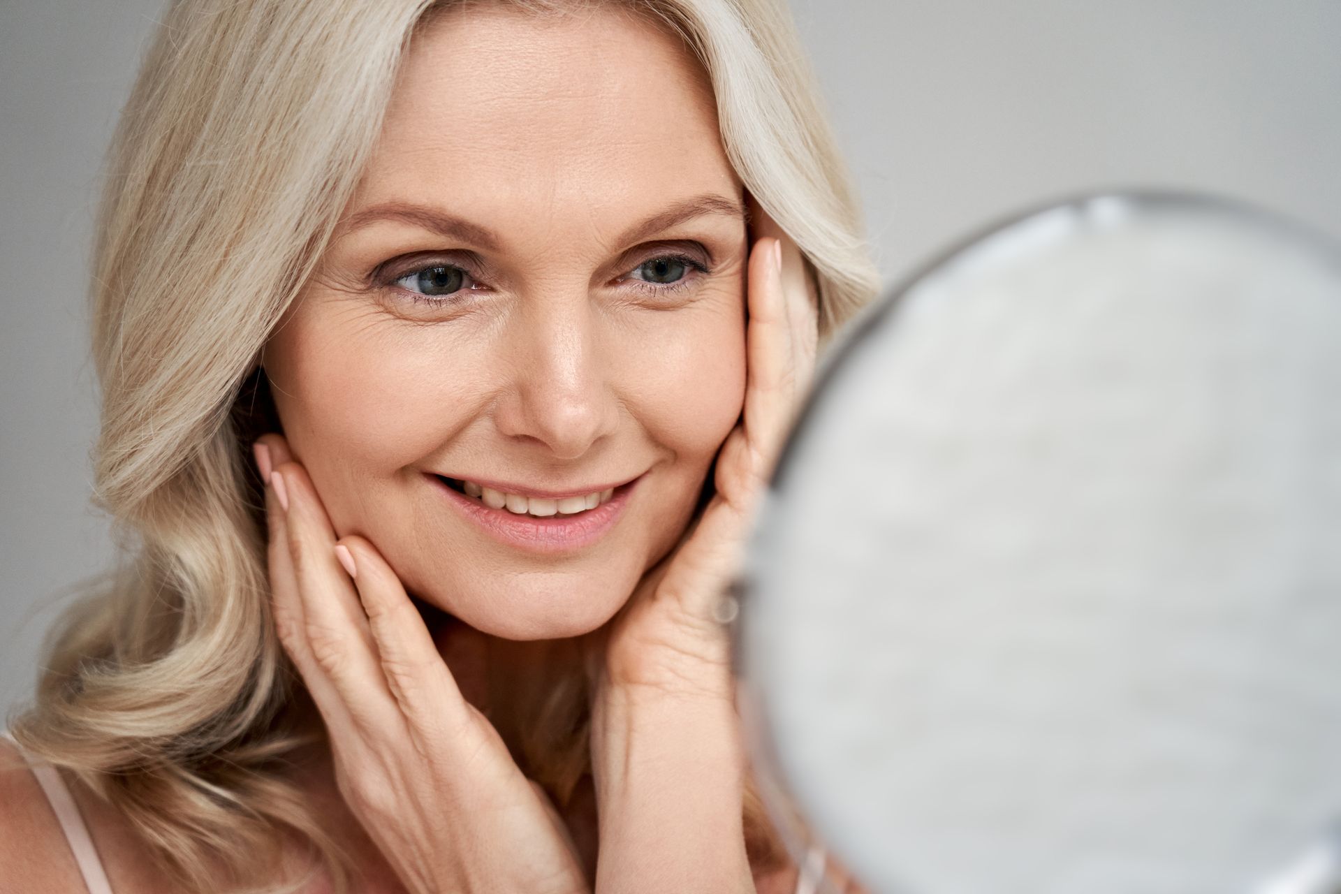 Woman with blonde hair smiling, looking in mirror, touching face.