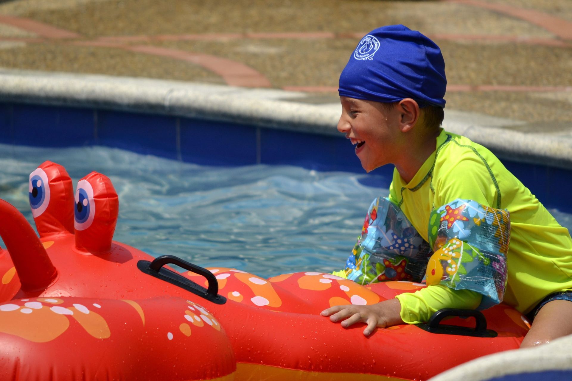 Niño en la piscina con gorro de natación y flotadores en los brazos, sonríe sentado sobre un cangrejo inflable rojo.