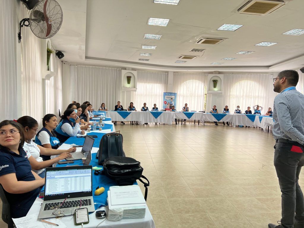 Un hombre se dirige a un grupo en una sala de conferencias. Las personas están sentadas alrededor de una mesa curva con ordenadores portátiles.