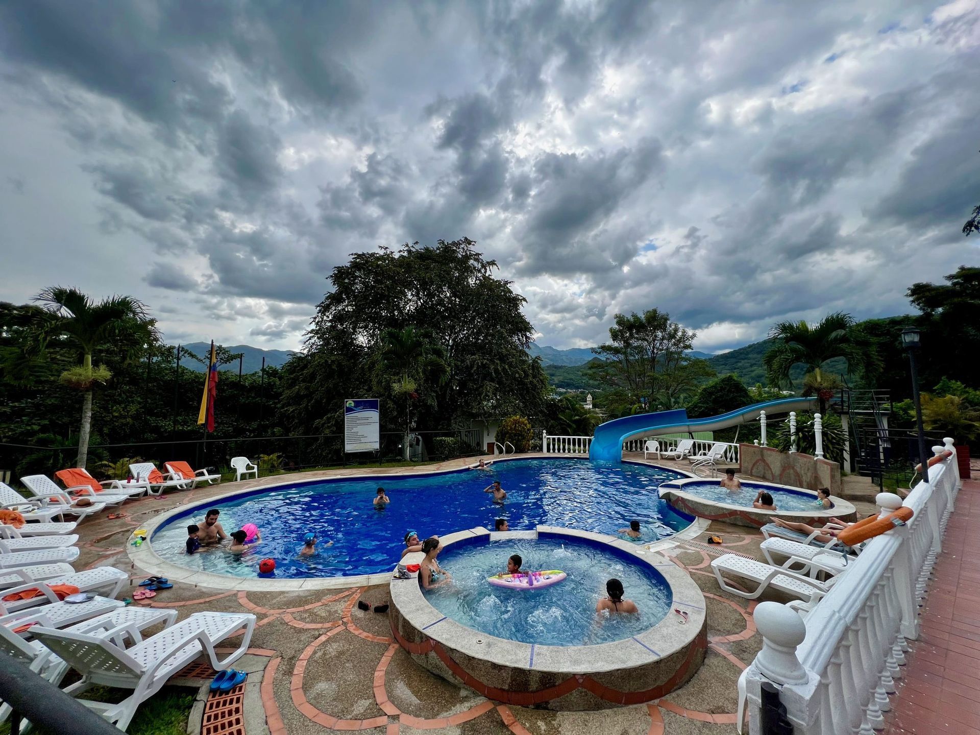 Piscina con gente, tobogán acuático y tumbonas bajo un cielo nublado.