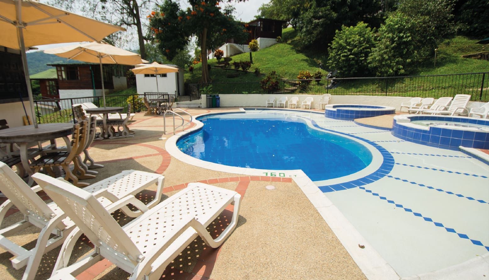 Piscina con tumbonas, mesas y jacuzzi, situada al pie de una colina cubierta de hierba.