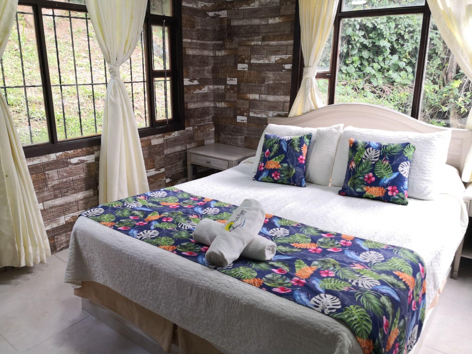 Dormitorio con cama, detalles florales, pared de ladrillo y ventanas con cortinas.