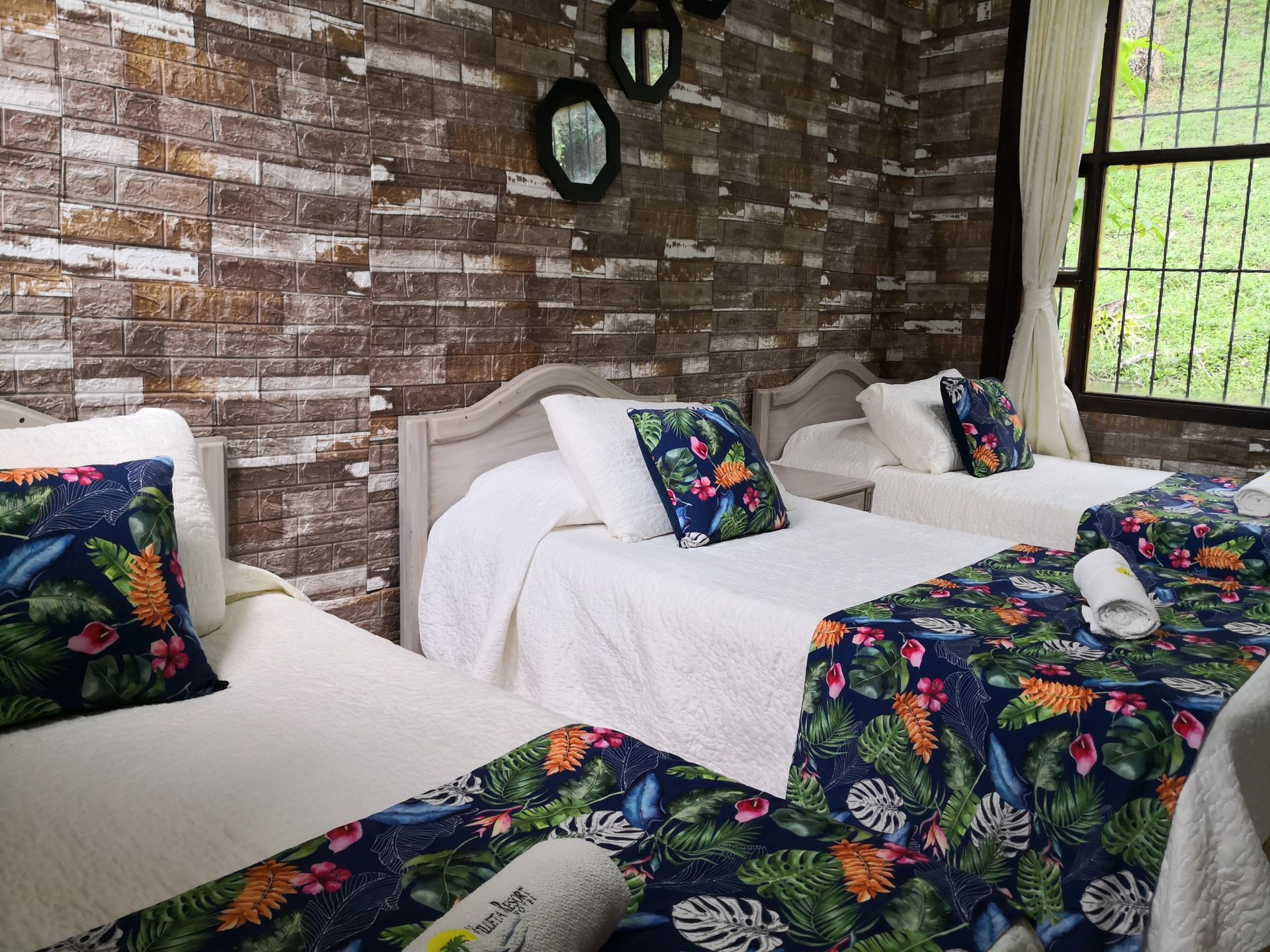 Dormitorio con tres camas; pared de ladrillo; cojines y caminos de cama con motivos florales; ventana.