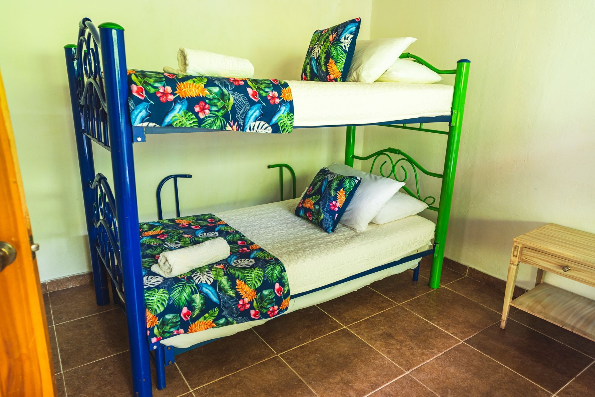 Litera con estructura azul y verde, ropa de cama con estampado tropical, almohadas blancas, en una habitación con suelo de baldosas.