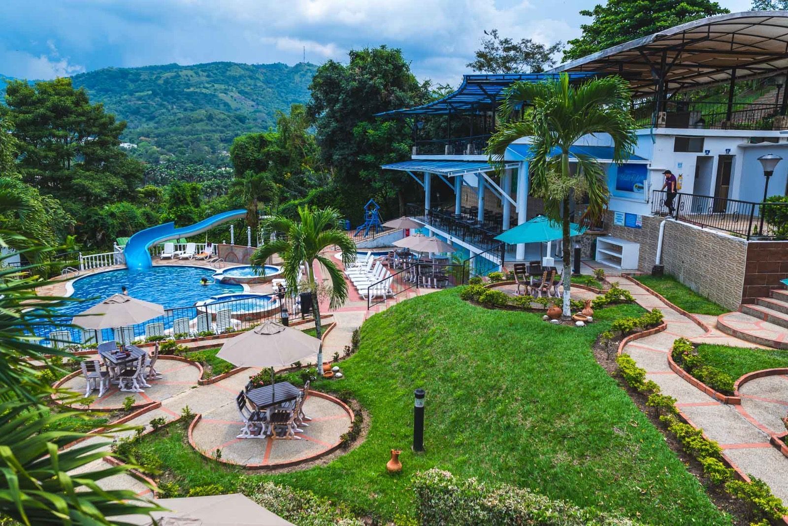 Vista aérea panorámica del complejo turístico con piscina, tobogán acuático y restaurante con vistas a una ladera boscosa.