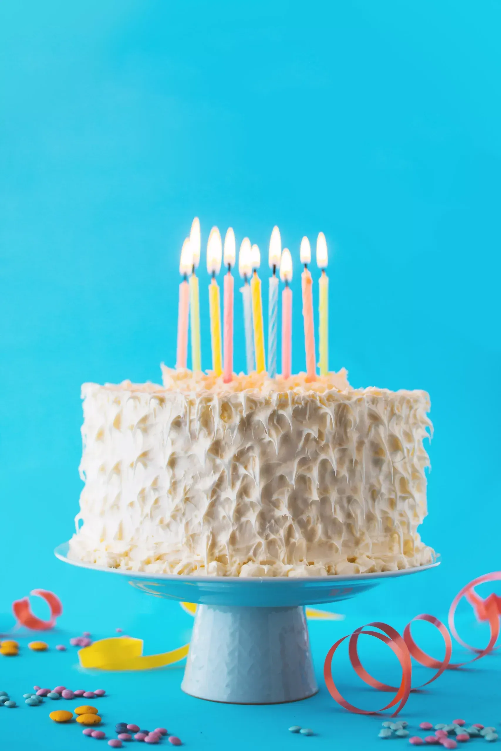 Tarta de cumpleaños con velas encendidas sobre un soporte azul, con fondo azul. Confeti.