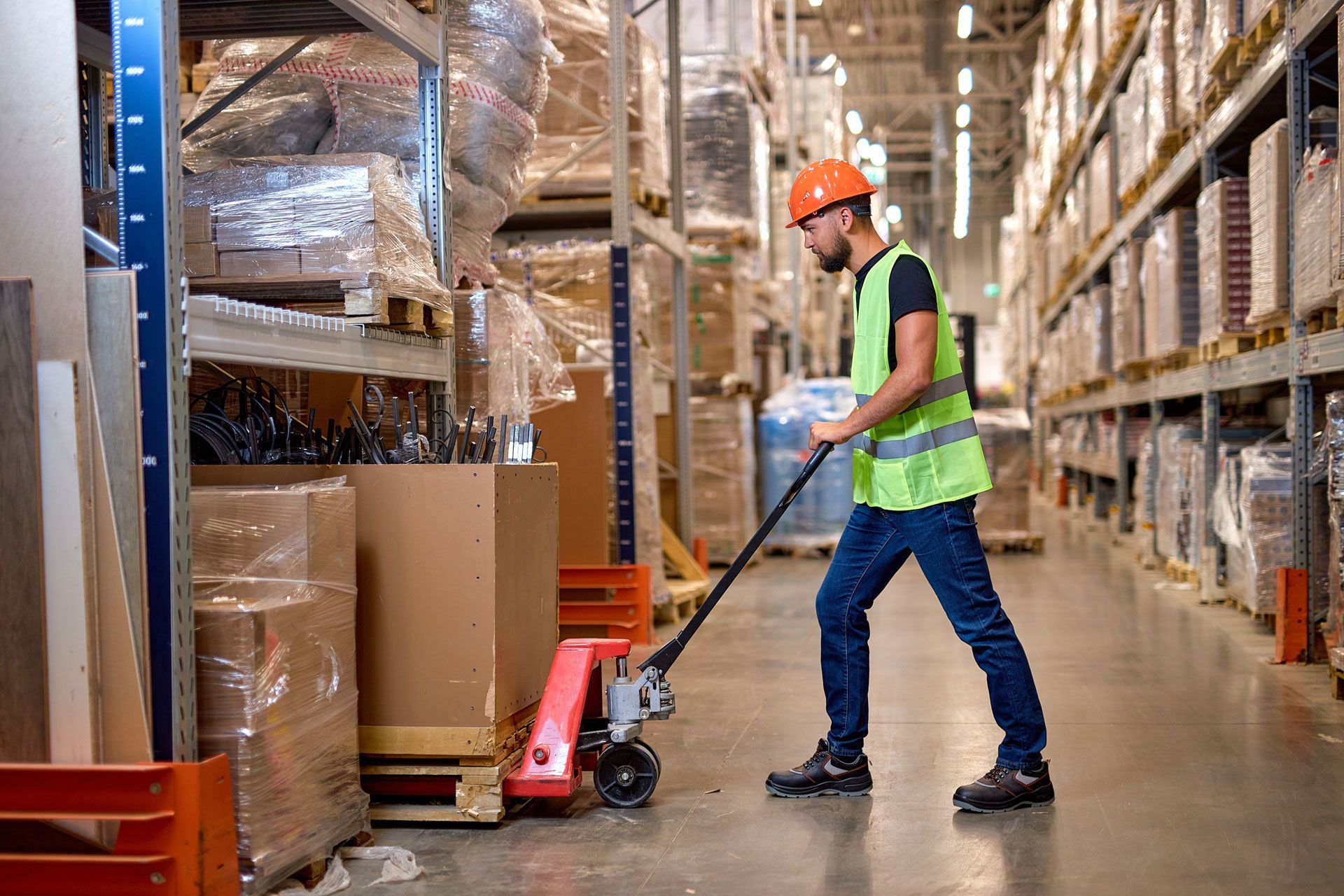 Warehouse worker in safety vest and hard hat pulling a pallet jack down a storage aisle.