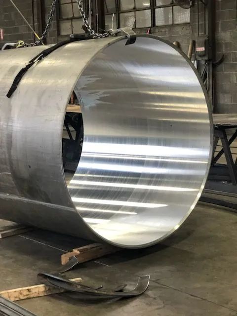 Large shiny metal cylindrical sheet in an industrial workshop, lying on the floor with tools nearby