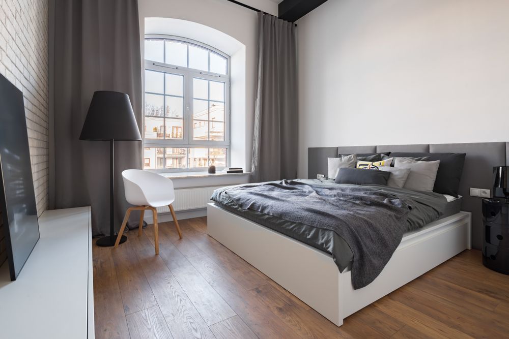 Bedroom with grey bedding, tall window with curtains, white brick wall, and hardwood floor.