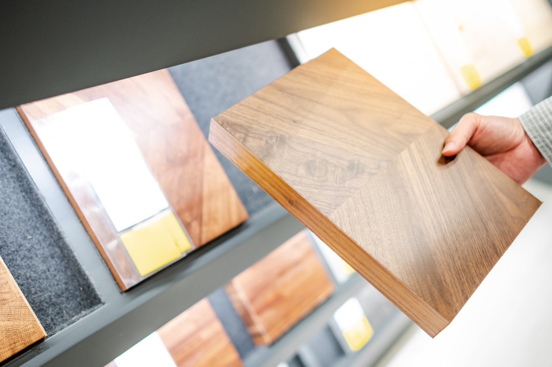 Hand holding a square wood sample, displaying it in front of a display of wood options.