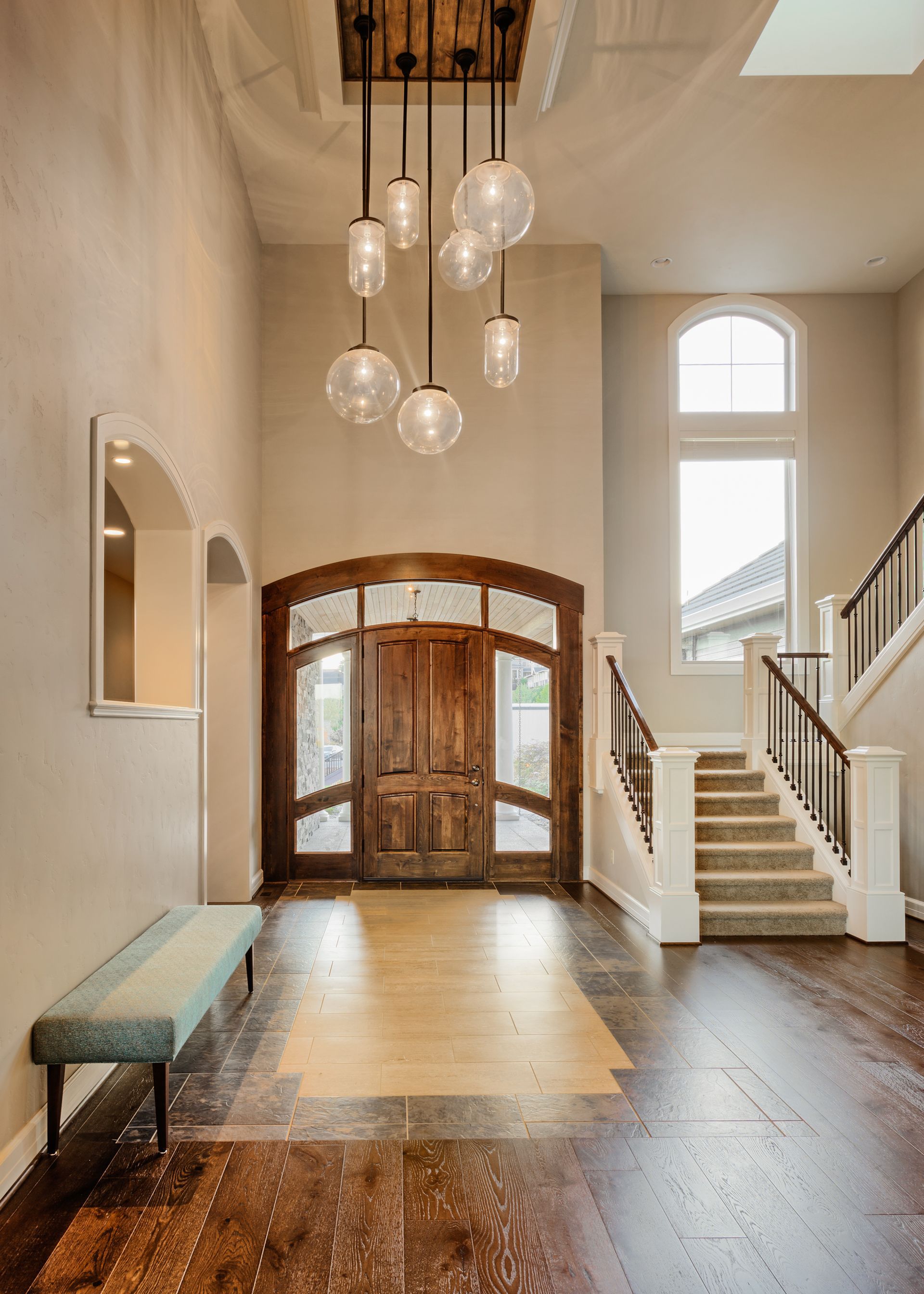Grand foyer with wood door, staircase, and light fixtures. Hardwood flooring, bench.