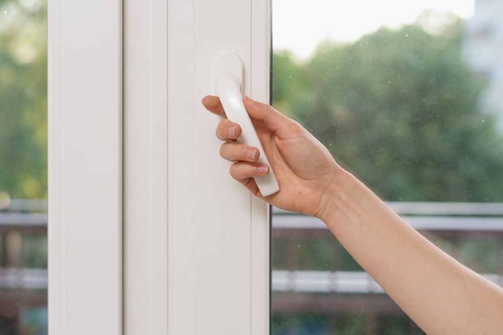Hand opening a white window, arm extended. Green trees visible through the glass.