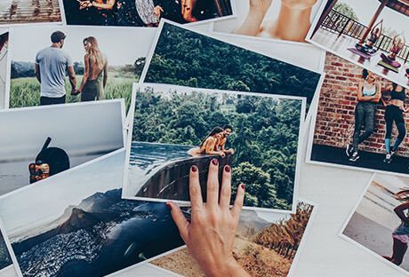 Scattered travel photos spread on a table, with one hand holding a photo of a car by a forested lake.