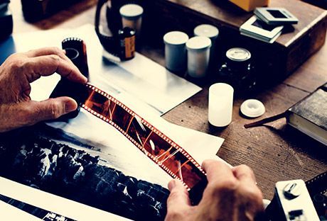 Hands holding a film negative over a table with photo prints, cups, and developing supplies