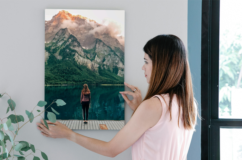 Woman adjusting a framed mountain photo on a white wall beside a window