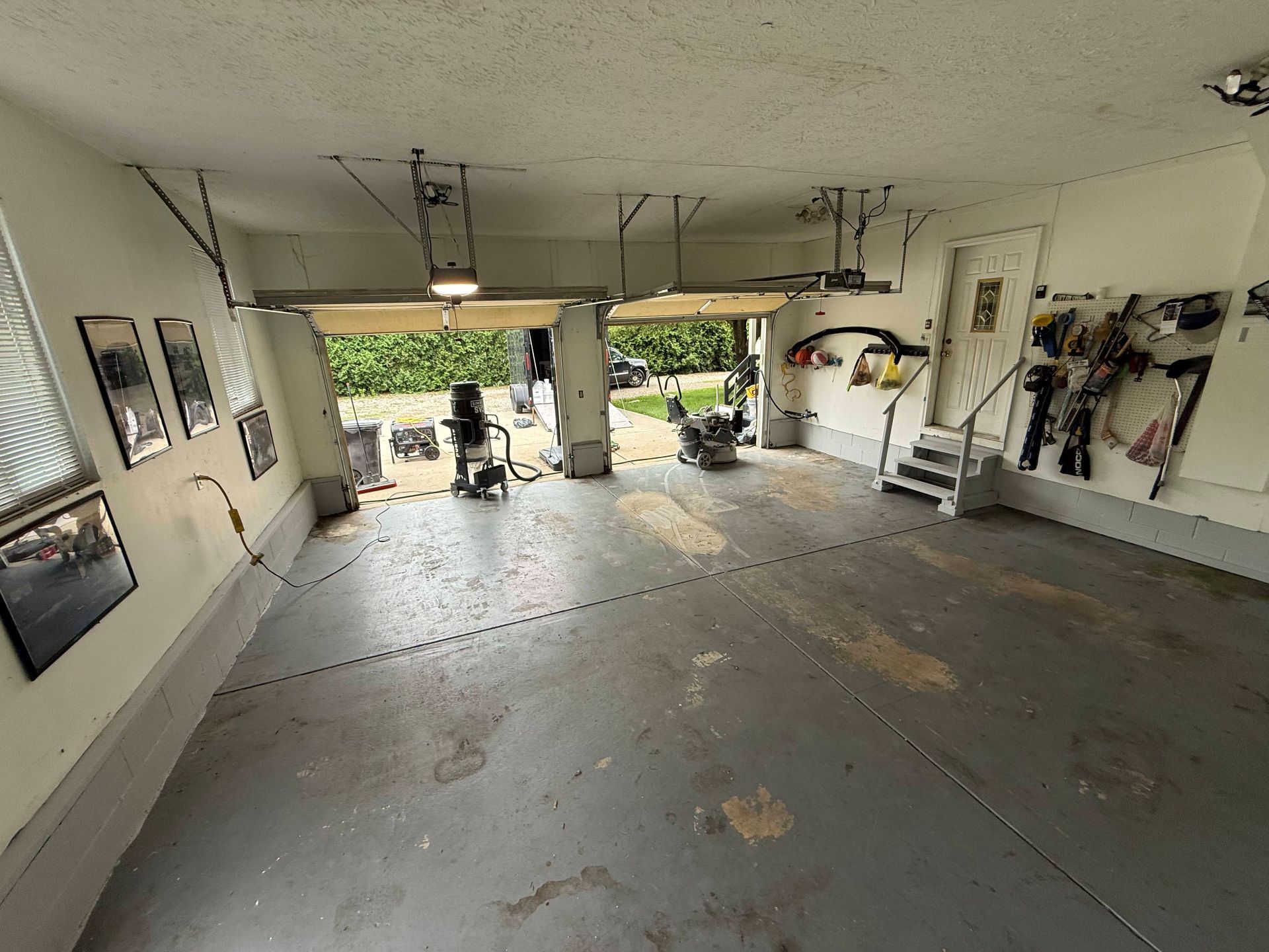 Empty garage with open door, gray floor, wall-mounted bikes and tools, and sunlight spilling in from outside
