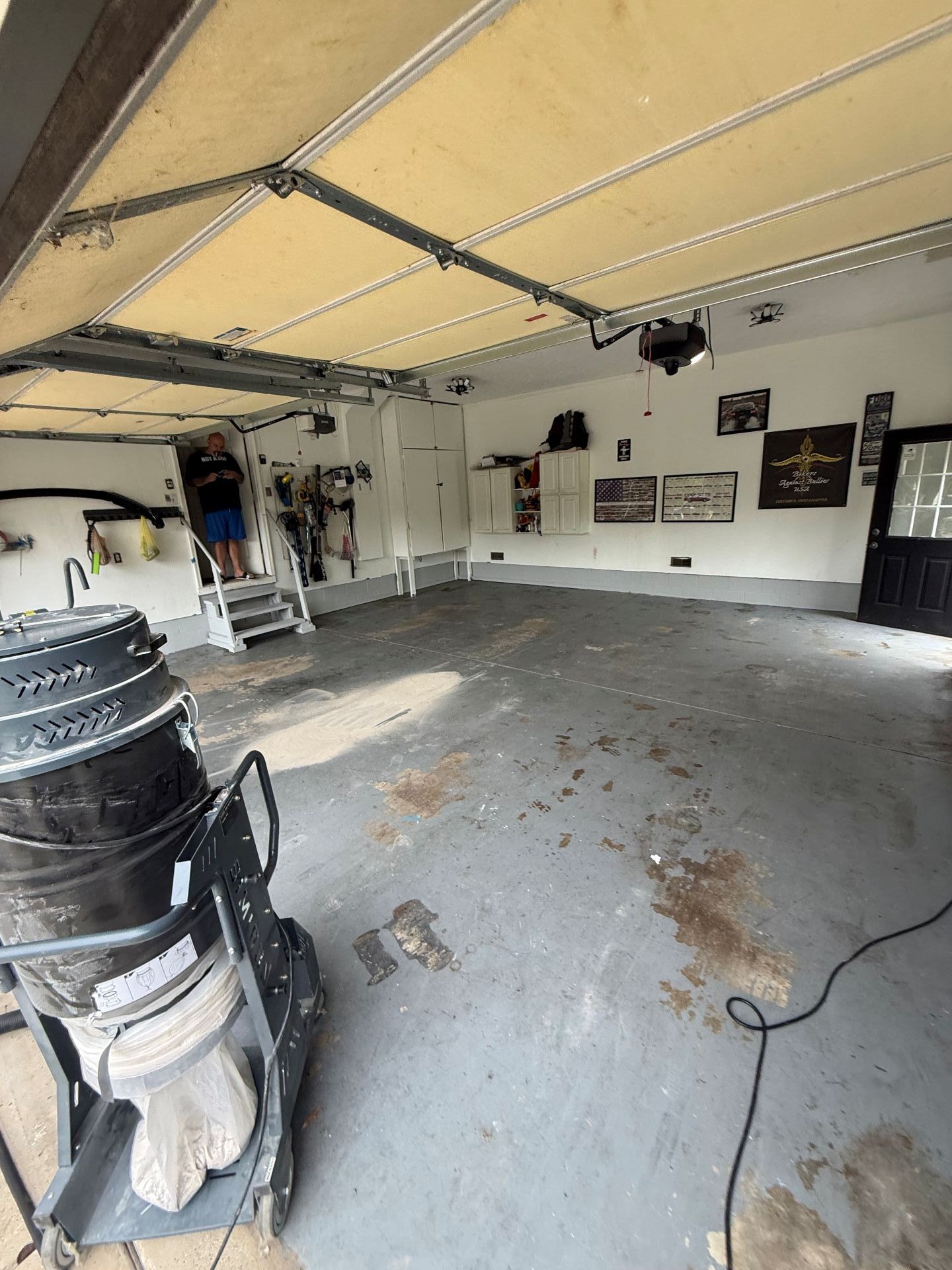 Empty garage with open overhead door, white walls, scattered items, and a concrete floor.