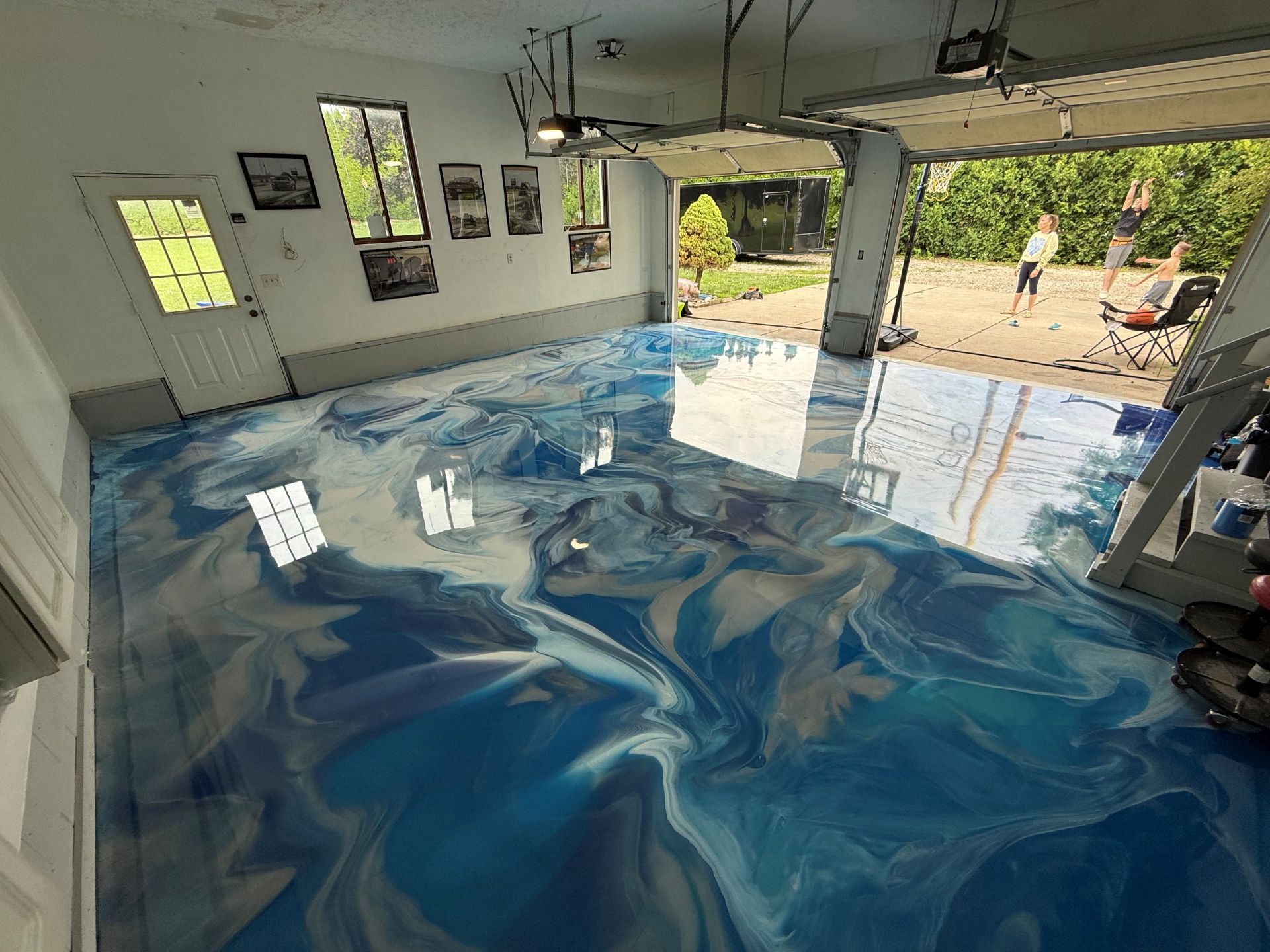Garage with a glossy blue-and-white epoxy floor, open to a sunny backyard patio and pool.