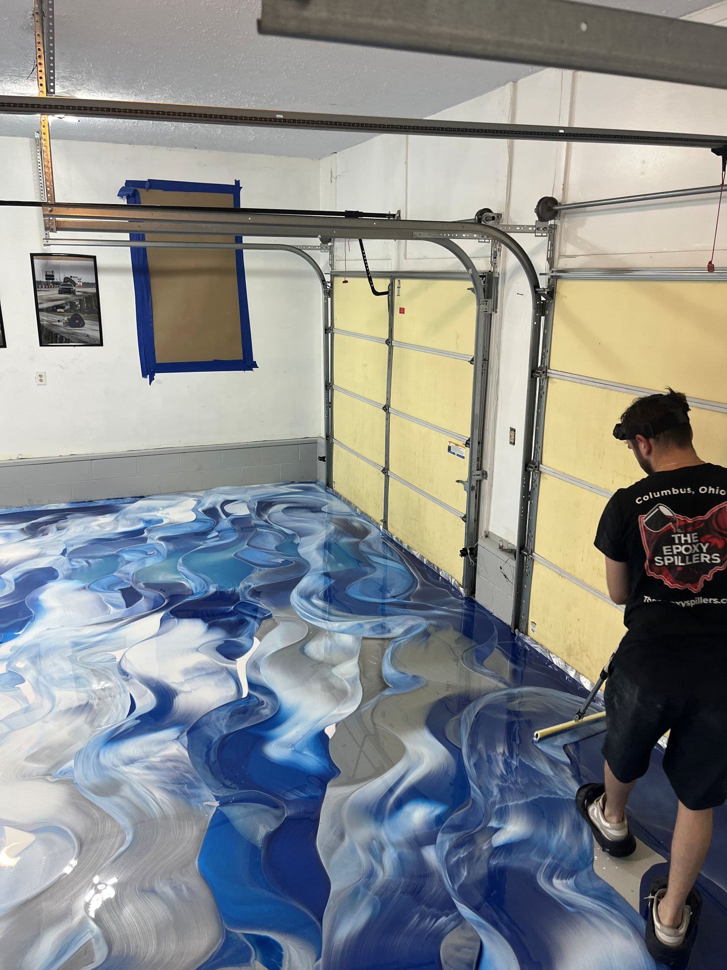 Garage interior with glossy blue-and-white marbled epoxy floor and a person painting near the open door.