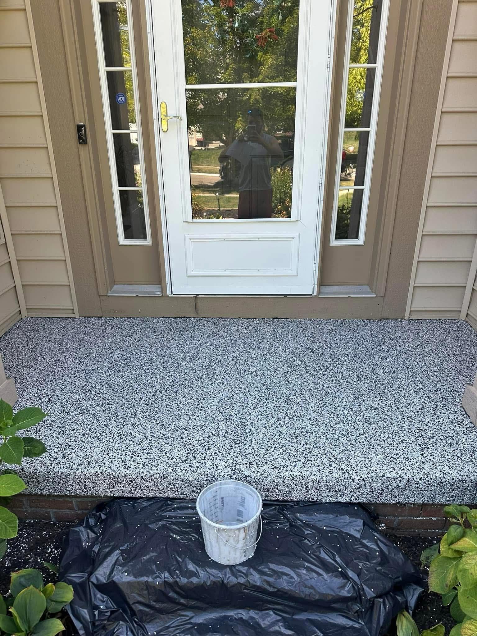 Front porch with speckled flooring, white door, shutters, and a small bucket on a black tarp by the steps