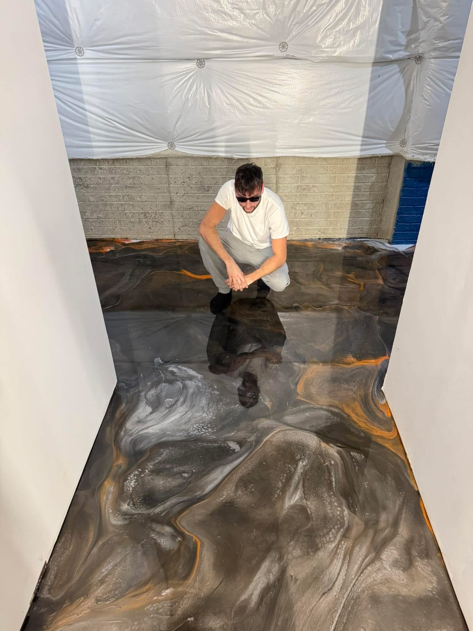 Person crouches over glossy black material in a room with plastic-covered walls and a gray floor.