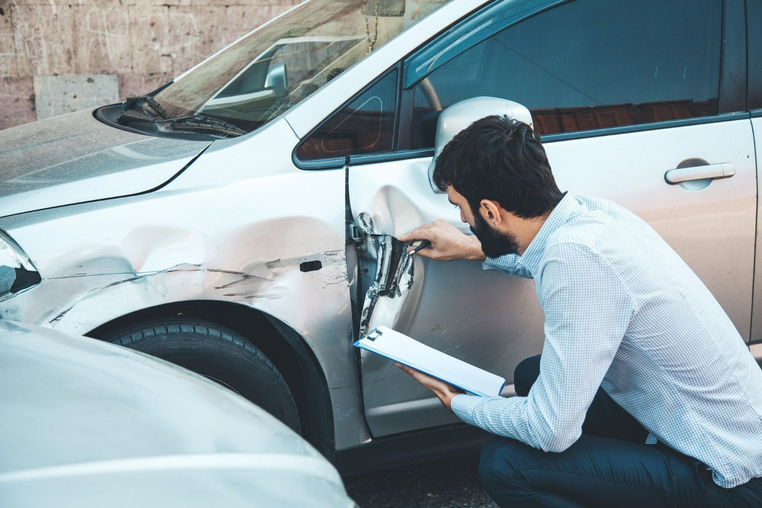 Man inspecting damage on the side of a silver car with a phone in hand and clipboard.