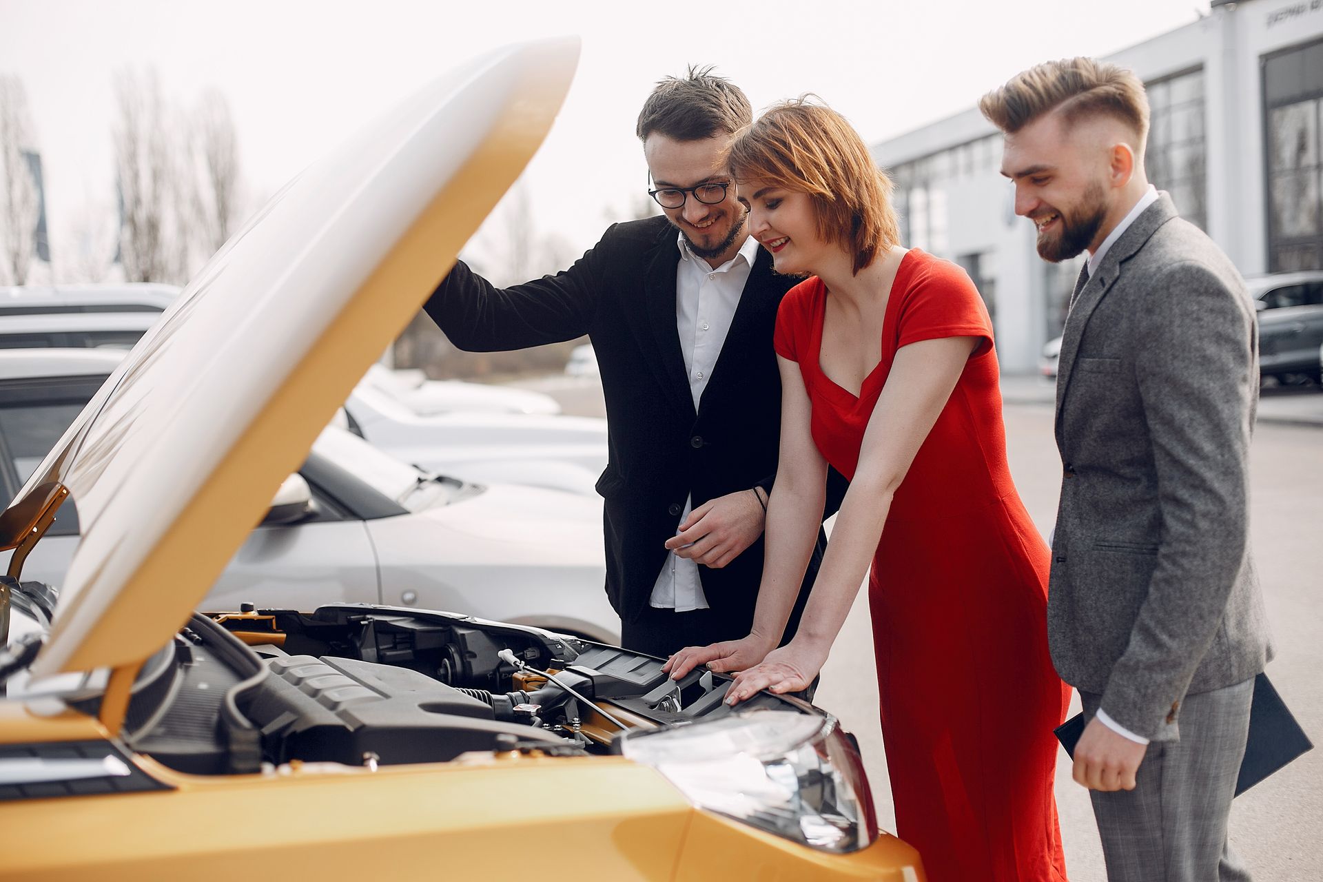 A man and two women are looking under the hood of a car.