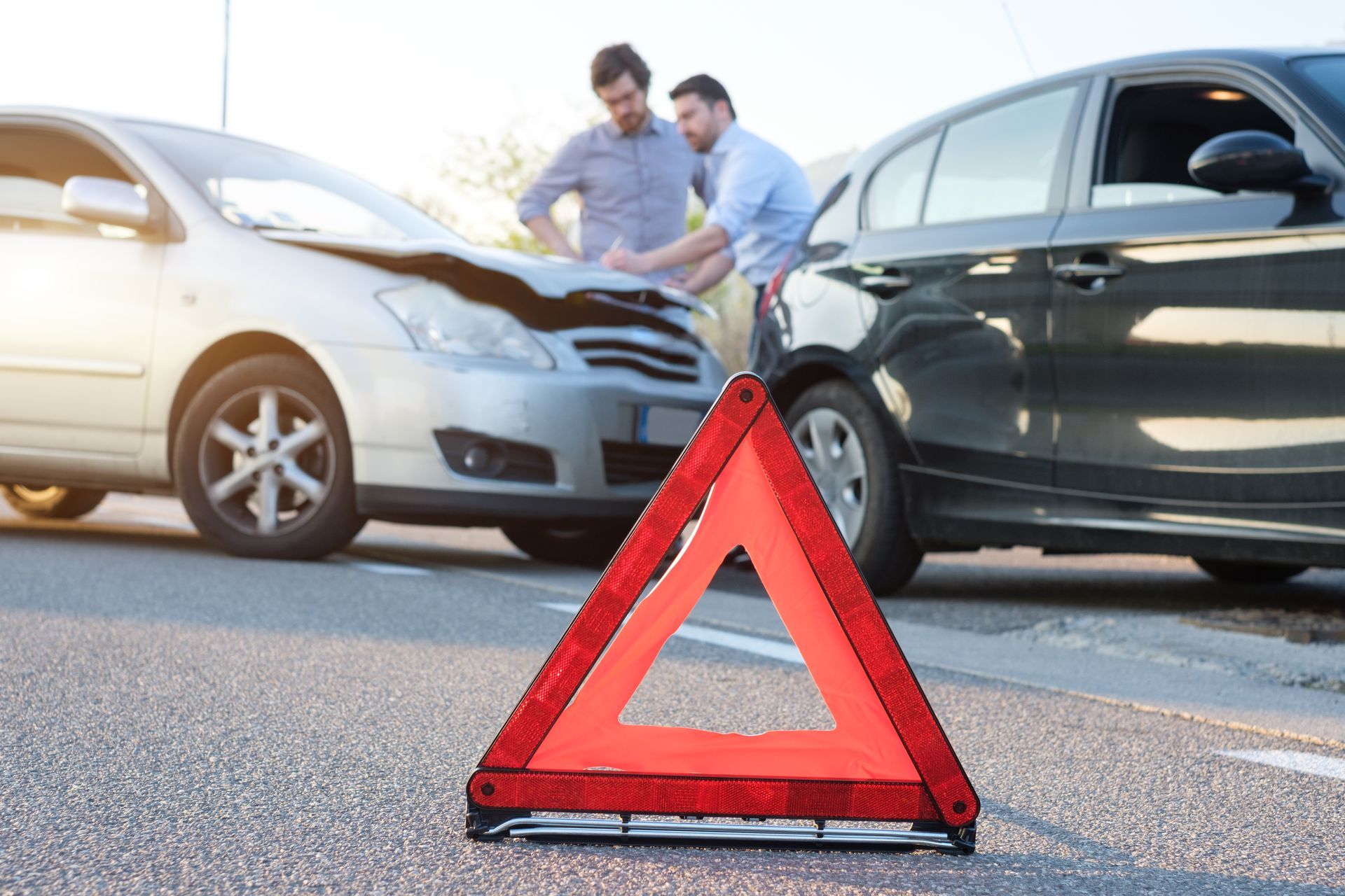 Two men inspect a car crash on a road, with a red warning triangle in the foreground.