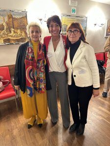 Tre donne sono in piedi insieme in un interno, in posa per una foto. Sono di fronte a un'opera d'arte, una con un abito giallo, una con pantaloni grigi e una con pantaloni neri.