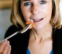 Donna che mangia con una forchetta, tenendo il cibo alla bocca. Ha i capelli biondi e indossa una giacca scura.