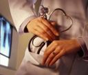 Medico con stetoscopio in mano, primo piano. Camice bianco, mani giunte, campana dello stetoscopio a forma di cuore. Radiografia sullo sfondo.