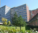 Vista esterna di edifici ospedalieri a più piani con alberi e vegetazione in primo piano sotto un cielo azzurro.