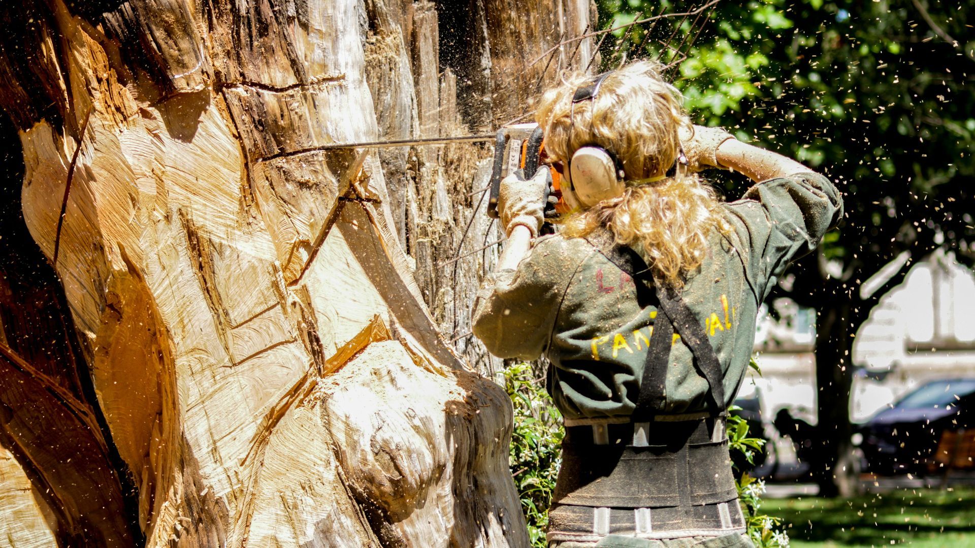 Person carving wood sculpture with a chainsaw, wearing safety gear outdoors, wood shavings flying.