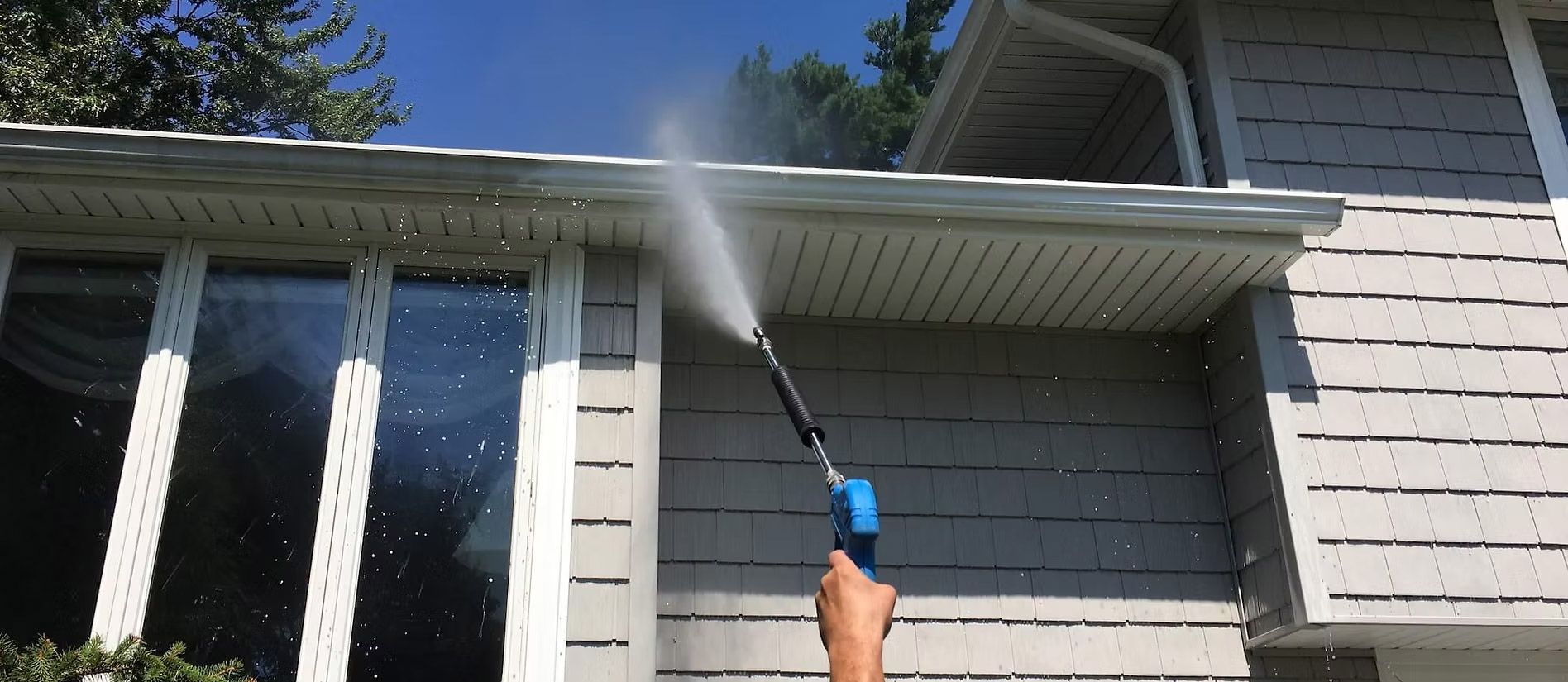 Person power washing the side of a house with a spray gun, outdoors on a sunny day.