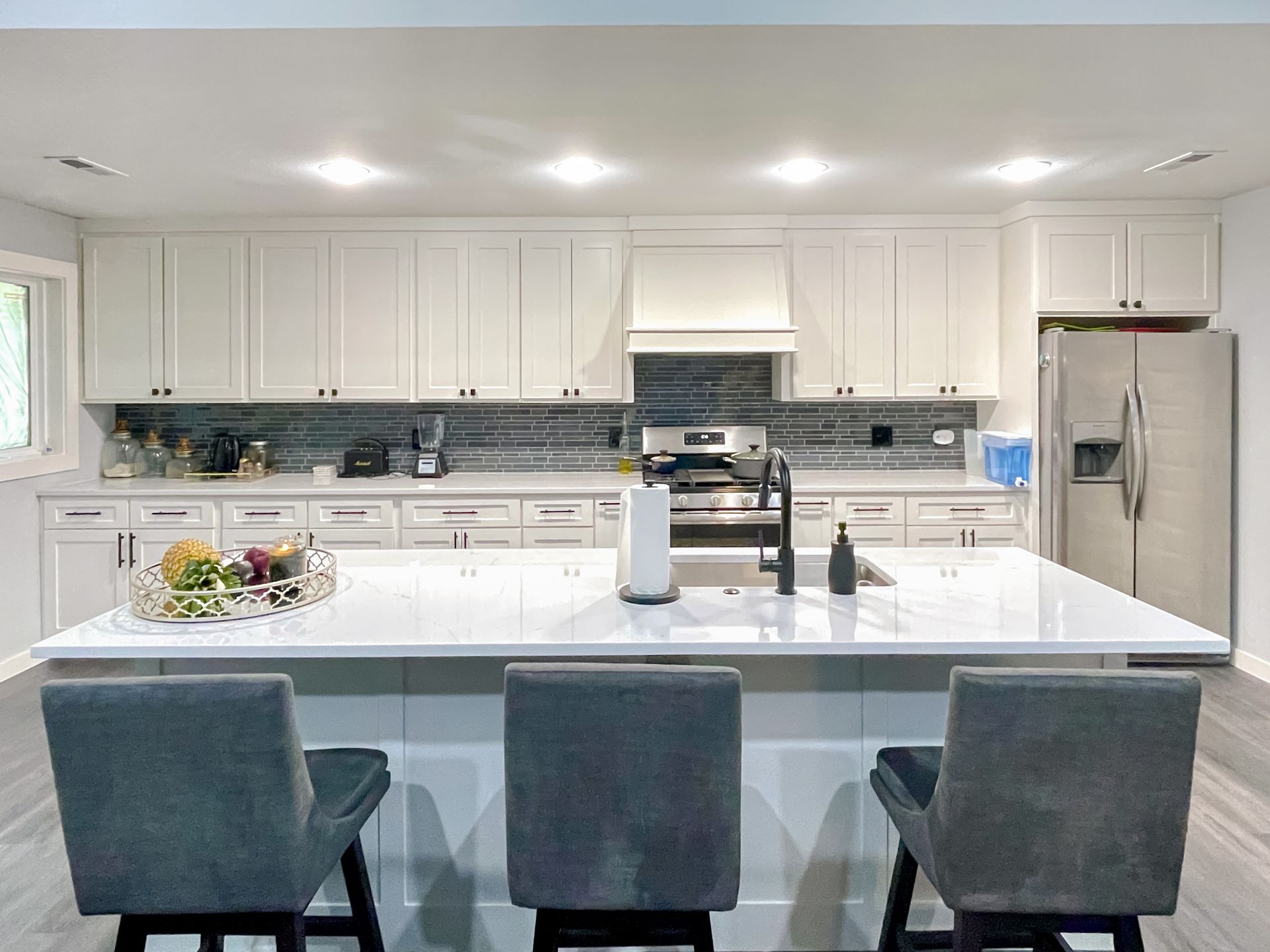 Modern white kitchen with island, gray bar stools, stainless steel appliances, and countertop.