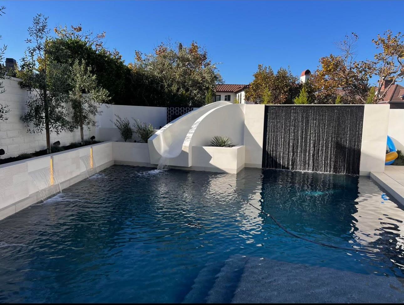 A pool with a slide and waterfall feature. Blue water, white slide, stone walls, and a sunny sky.