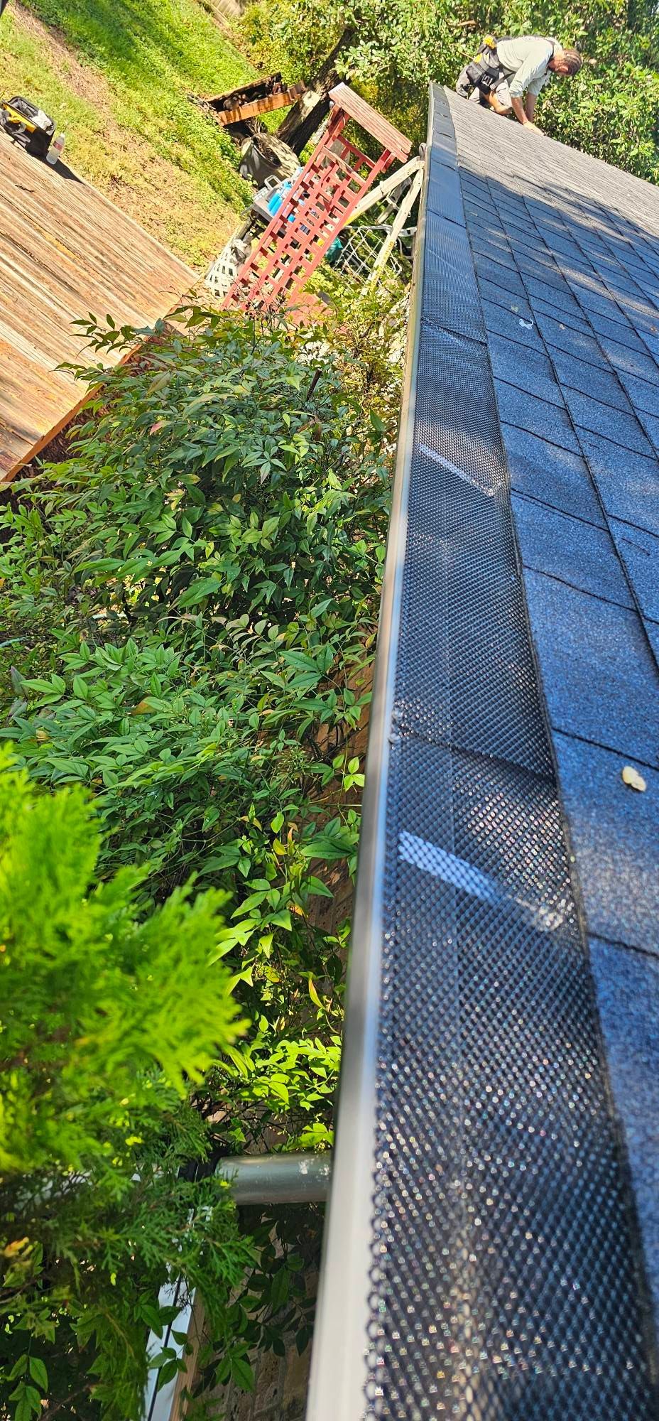 A close-up, high-angle view of a mesh gutter guard installed along the edge of a roof, next to green foliage.